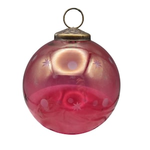 May include: A vintage, round Christmas ornament in shades of pink and red. The glass ornament features etched star and dot patterns. It has a brass-colored metal cap and a loop for hanging.