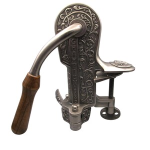 Puede incluir: Un pelador de manzanas Champion 1897 de metal plateado con un patrón floral en relieve en el cuerpo principal. El dispositivo cuenta con un mango de metal curvado con una empuñadura de madera marrón oscuro, una abrazadera de tornillo y un brazo mecánico.