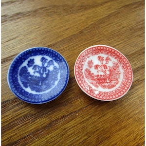 May include: Two miniature decorative plates. One is blue with a white floral design, and the other is red with a white landscape scene. Both plates have a detailed border and are set on a wooden surface.