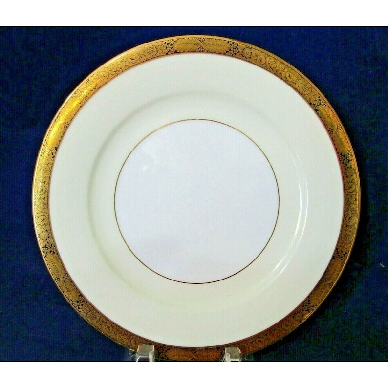 Op de afbeelding: Een wit keramisch dinerbord met een gouden rand en een ingewikkeld gouden ontwerp. Het bord heeft een licht verhoogd midden.