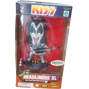 Puede incluir: Una figura de Gene Simmons Rock Headliners XL en una caja roja, blanca y negra. La figura es una caricatura de Gene Simmons, el bajista de la banda de rock KISS. La figura sostiene un bajo y tiene la lengua larga fuera. La caja tiene las palabras "Rock Headliners XL" y "Gene Simmons" en ella.