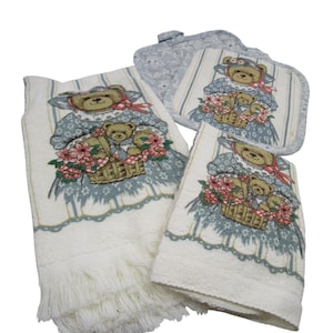 May include: A collection of kitchen textiles with a teddy bear motif. The set comprises a fringed towel, a hand towel, a pot holder, and a smaller towel. Each piece showcases a teddy bear in a dress, carrying a basket of flowers. The color palette is white, blue, and beige.