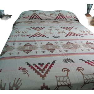 Puede incluir: Una colcha crema con un diseño del suroeste. El patrón incluye formas geométricas, ciervos y símbolos solares en tonos marrones, rojos y beige. La colcha está extendida sobre una cama.