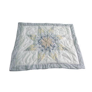 May include: A square, quilted pillow with a starburst design. The star is made of various pastel fabrics, including yellow, blue, and pink. The pillow has a white background and a blue and white gingham border.