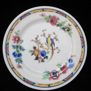 May include: White ceramic plate with a colourful floral and bird design. The plate features a central image of a bird perched on a flowering branch, surrounded by a border of flowers and a yellow and blue trim.