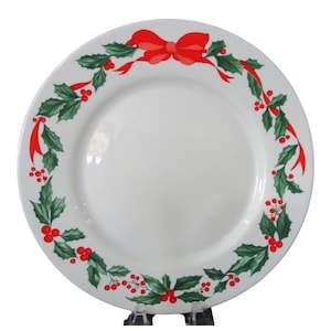 May include: White ceramic dinner plate with a green holly and red berry border and a red bow.