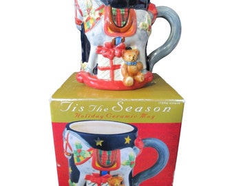 Pode incluir: Uma caneca de cerâmica de Natal com um design de cavalo de balanço. A caneca é decorada com um cavalo de balanço vermelho, branco e azul, um urso de peluche e presentes. A caneca está numa caixa com as palavras "Tis The Season Holiday Ceramic Mug" impressas nela.