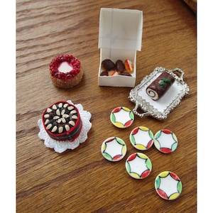 May include: A collection of miniature food items, including a small cake with red icing, a cake with nuts, a box of chocolates, a chocolate roll on a silver tray, and several small, colourful plates. The items are on a wooden surface.