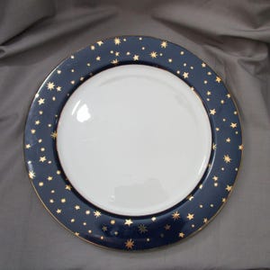 May include: A white ceramic plate with a navy blue rim featuring gold star detailing. The plate has a gold edge. This plate is designed for serving food, with a diameter of approximately 25 cm.