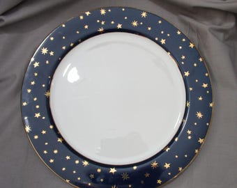 May include: A round, white ceramic plate with a navy blue rim decorated with gold stars. The plate has a gold trim around the edge. The plate is designed for serving food.