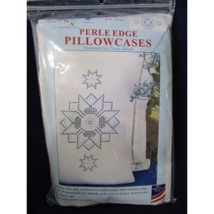 Puede incluir: Un paquete que contiene dos fundas de almohada blancas con un diseño de estrella azul y blanca, estampadas para punto de cruz. El paquete está etiquetado como "Perle Edge Pillowcases" y "XX Stars".