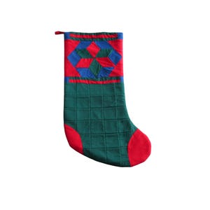 May include: A quilted Christmas stocking in green, red, and blue. The toe and top are red, with a geometric design in red, blue, and green on the upper portion. The main body is green with a quilted pattern.