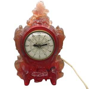 May include: A vintage red and amber-colored electric clock with a decorative, ornate design. The clock face is white with black numerals and hands. The clock has a power cord attached.