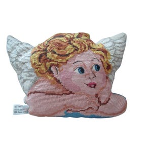 May include: A needlepoint cushion shaped like a cherub with curly blonde hair, blue eyes, and white wings. The figure has pink skin and is resting on a blue base. A small white tag on the side reads: NOT A TOY, House of Lloyd, Inc., Made in China, 100% Polyester Stuffing.