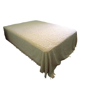 May include: A cream-coloured bedspread with a textured floral pattern and fringe trim. The bed covering is rectangular and covers the bed. The fringe hangs down from the edges of the bedspread. The bedspread is made of a soft material.