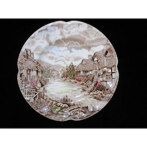 Puede incluir: Un plato de cerámica decorativo con una detallada escena de paisaje de un pueblo con casas con tejados de paja, un río y cielos nublados. El plato tiene un borde festoneado y es principalmente marrón, beige y azul.