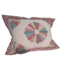 Può includere: Un cuscino trapuntato rettangolare con un motivo centrale a ventaglio in tonalità pastello di rosa, blu, giallo e stampe floreali. Il cuscino presenta un bordo arricciato con un motivo floreale su fondo rosa e tessuto bianco.