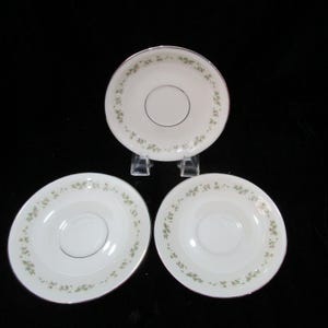 Könnte beinhalten: Drei weiße Untertassen mit einem zarten Blumenmuster in Grün und Rosa am Rand. Jede Untertasse hat eine silberfarbene Umrandung und eine zentrale Vertiefung. Die Untertassen werden auf einer schwarzen Oberfläche präsentiert.