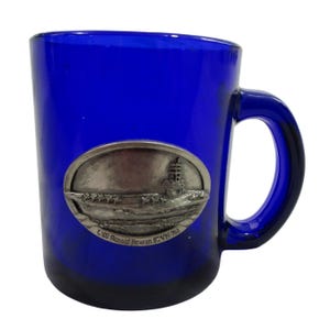 May include: A blue glass mug with a pewter emblem of the USS Ronald Reagan (CVN-76) aircraft carrier. The emblem is oval shaped and features a detailed image of the ship.
