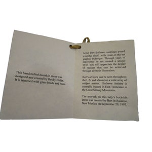 Puede incluir: Un primer plano de una tarjeta doblada con texto que describe un vestido de piel de venado hecho a mano. El vestido fue dise&ntilde;ado y creado por Becky Nufer y adornado con cuentas de vidrio y hueso. La tarjeta tambi&eacute;n describe al artista, Bert Ballowe, y su estilo de ilustraci&oacute;n con aer&oacute;grafo. La obra de arte fue creada en Ruidoso, Nuevo M&eacute;xico, el 20 de septiembre de 1997.