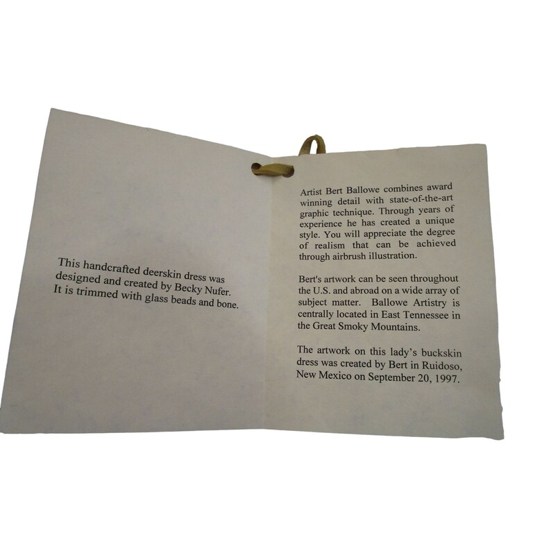 Puede incluir: Un primer plano de una tarjeta doblada con texto que describe un vestido de piel de venado hecho a mano. El vestido fue dise&ntilde;ado y creado por Becky Nufer y adornado con cuentas de vidrio y hueso. La tarjeta tambi&eacute;n describe al artista, Bert Ballowe, y su estilo de ilustraci&oacute;n con aer&oacute;grafo. La obra de arte fue creada en Ruidoso, Nuevo M&eacute;xico, el 20 de septiembre de 1997.
