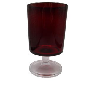 May include: A burgundy-colored glass goblet with a clear, circular base and stem. The glass has a slightly textured appearance. The goblet is designed to hold liquids and is suitable for beverages.