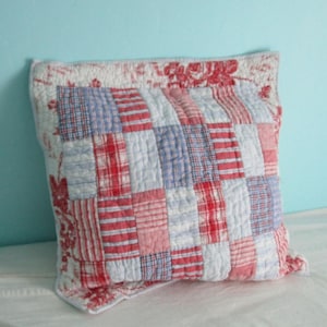 May include: A square quilted throw pillow featuring a central patchwork design of red, white, and blue fabric rectangles in various plaid and striped patterns. The border of the pillow displays a red and white floral toile print on a white background.