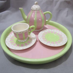 May include: A pastel-colored ceramic tea set with a pink and green striped teapot, cup, saucer, and plate. The set is arranged on a pink tray with a green rim. The cup and saucer have a matching design.