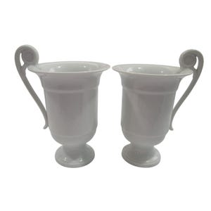 May include: Two white ceramic cups with ornate handles and a pedestal base. The cups are empty and ready to be filled with a beverage.