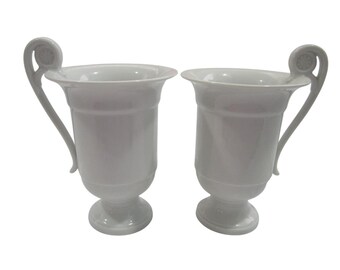 Puede incluir: Dos tazas de cerámica blanca con asas ornamentadas y una base de pedestal. Las tazas están vacías y listas para llenarse con una bebida.