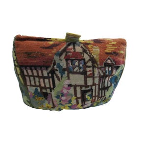 May include: A vintage needlepoint bag featuring a detailed scene of a Tudor-style house with a red roof and brown timber framing. The bag's design includes colorful floral and landscape elements, with a small handle at the top.