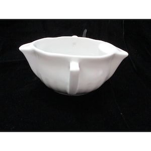 Puede incluir: Una salsera de porcelana blanca con asa y boquilla. La salsera tiene un cuerpo redondeado y un acabado liso y brillante. El asa es rectangular y está unida al lateral. La boquilla está en el lado opuesto al asa.