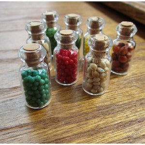 May include: A selection of small glass bottles with cork stoppers, each filled with colourful beads. The beads are various colours, including green, red, yellow, and beige. The bottles are arranged on a wooden surface.