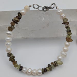 K&ouml;nnte beinhalten: Ein Armband aus wei&szlig;en Perlen und gr&uuml;nen und braunen Edelsteinperlen. Das Armband hat einen silbernen Verschluss.