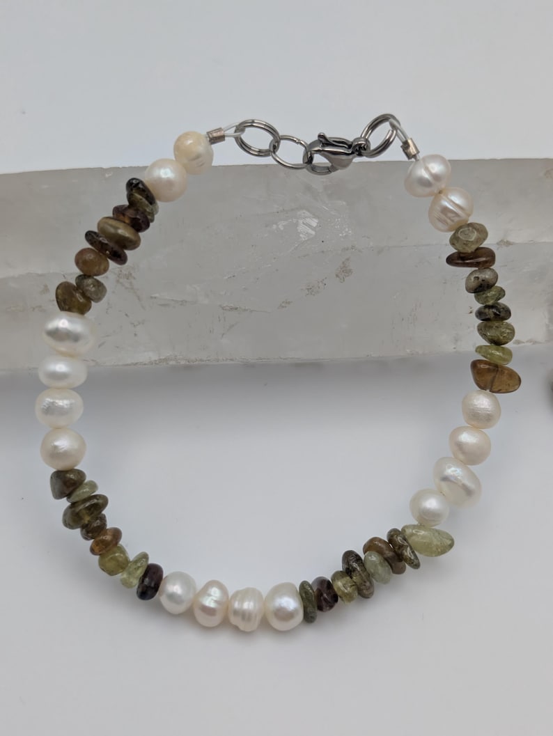 K&ouml;nnte beinhalten: Ein Armband aus wei&szlig;en Perlen und gr&uuml;nen und braunen Edelsteinperlen. Das Armband hat einen silbernen Verschluss.