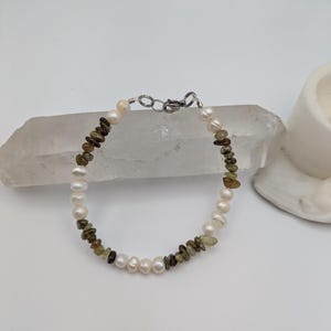 K&ouml;nnte beinhalten: Ein zartes Armband mit abwechselnden wei&szlig;en Perlen und gr&uuml;nen und braunen Edelsteinperlen. Das Armband wird mit einem silbernen Verschluss befestigt.