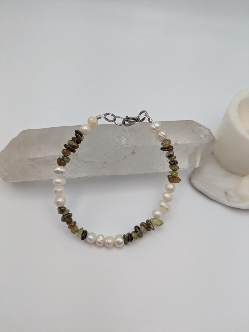 K&ouml;nnte beinhalten: Ein zartes Armband mit abwechselnden wei&szlig;en Perlen und gr&uuml;nen und braunen Edelsteinperlen. Das Armband wird mit einem silbernen Verschluss befestigt.