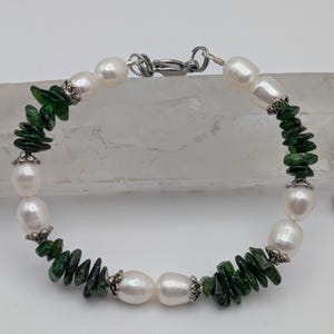 Op de afbeelding: Een armband gemaakt met witte parels en groene edelsteen chips. De armband heeft een zilveren sluiting.