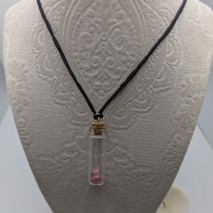 Puede incluir: Un collar de cord&oacute;n negro con un peque&ntilde;o colgante de frasco de vidrio que contiene cristales rosas. El frasco tiene un tap&oacute;n de corcho.