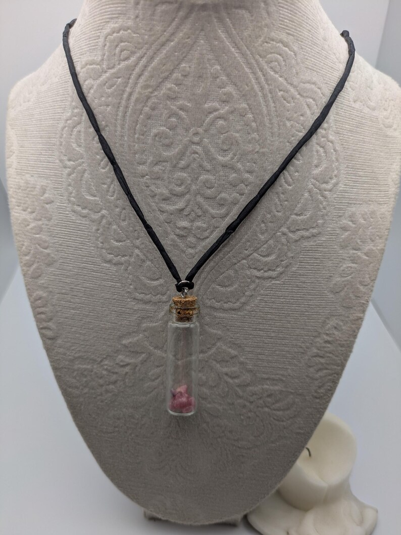 Puede incluir: Un collar de cord&oacute;n negro con un peque&ntilde;o colgante de frasco de vidrio que contiene cristales rosas. El frasco tiene un tap&oacute;n de corcho.