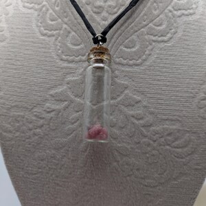 Puede incluir: Un collar con un vial de vidrio y un cord&oacute;n negro. El vial contiene peque&ntilde;os cristales rosas.