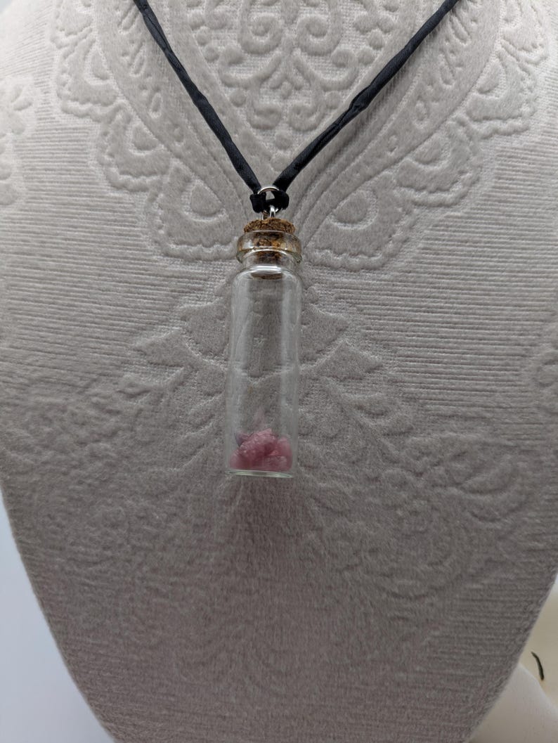 Puede incluir: Un collar con un vial de vidrio y un cord&oacute;n negro. El vial contiene peque&ntilde;os cristales rosas.