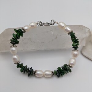 Op de afbeelding: Een armband van witte parels en groene edelstenen met een zilveren sluiting. De armband is gemaakt met afwisselende witte parels en groene edelstenen.