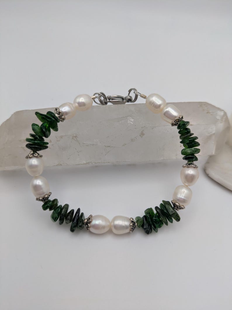 Op de afbeelding: Een armband van witte parels en groene edelstenen met een zilveren sluiting. De armband is gemaakt met afwisselende witte parels en groene edelstenen.