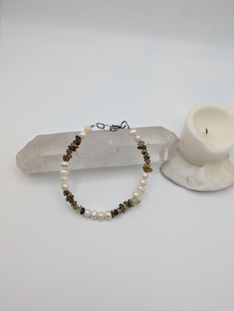 K&ouml;nnte beinhalten: Ein zartes Armband aus wei&szlig;en Perlen und gr&uuml;nen Edelsteinen. Das Armband liegt auf einer wei&szlig;en Oberfl&auml;che mit einer wei&szlig;en Kerze im Hintergrund.
