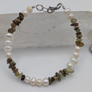 K&ouml;nnte beinhalten: Ein Armband aus wei&szlig;en Perlen und gr&uuml;nen und braunen Edelsteinperlen. Das Armband hat einen silbernen Verschluss.