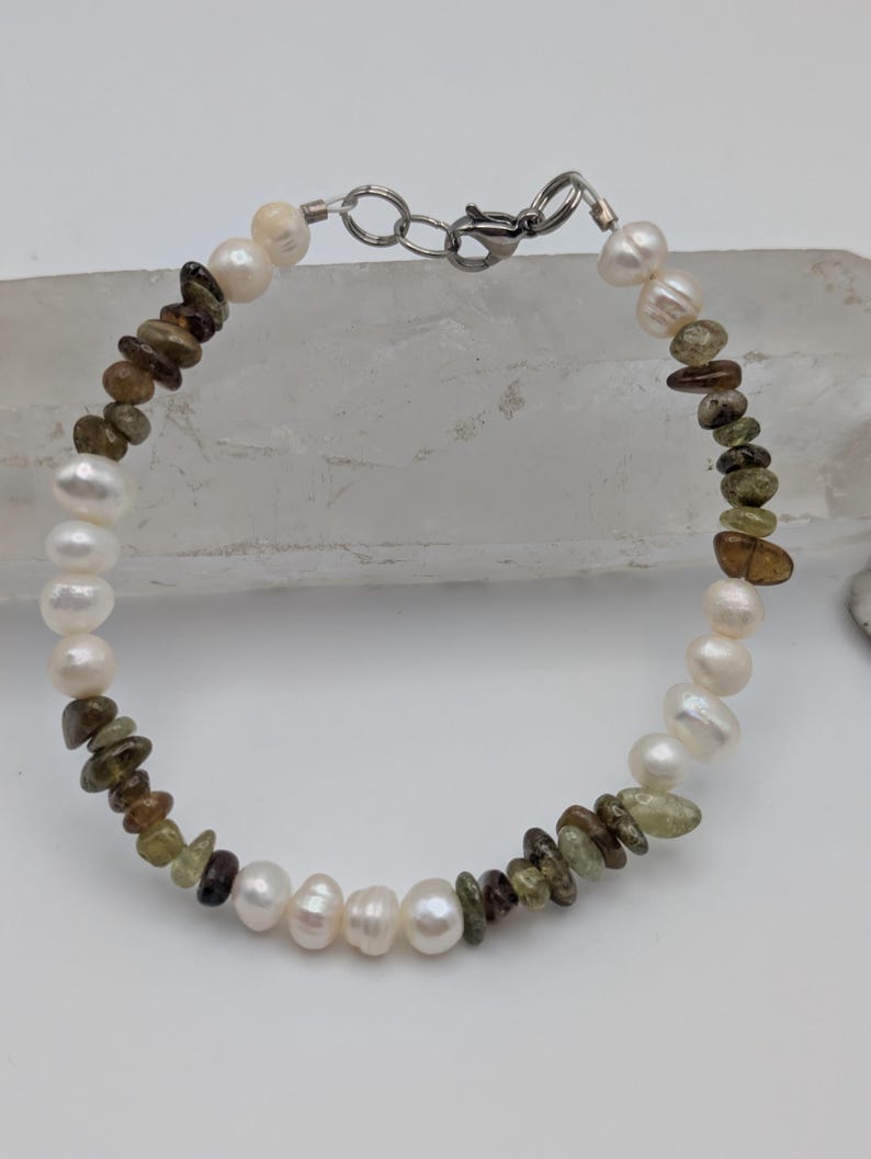 K&ouml;nnte beinhalten: Ein Armband aus wei&szlig;en Perlen und gr&uuml;nen und braunen Edelsteinperlen. Das Armband hat einen silbernen Verschluss.