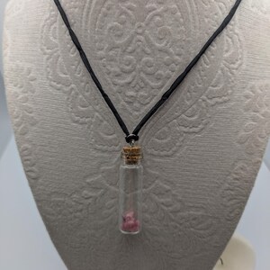 Puede incluir: Un collar de cord&oacute;n negro con un peque&ntilde;o colgante de frasco de vidrio que contiene cristales rosas. El frasco tiene un tap&oacute;n de corcho.