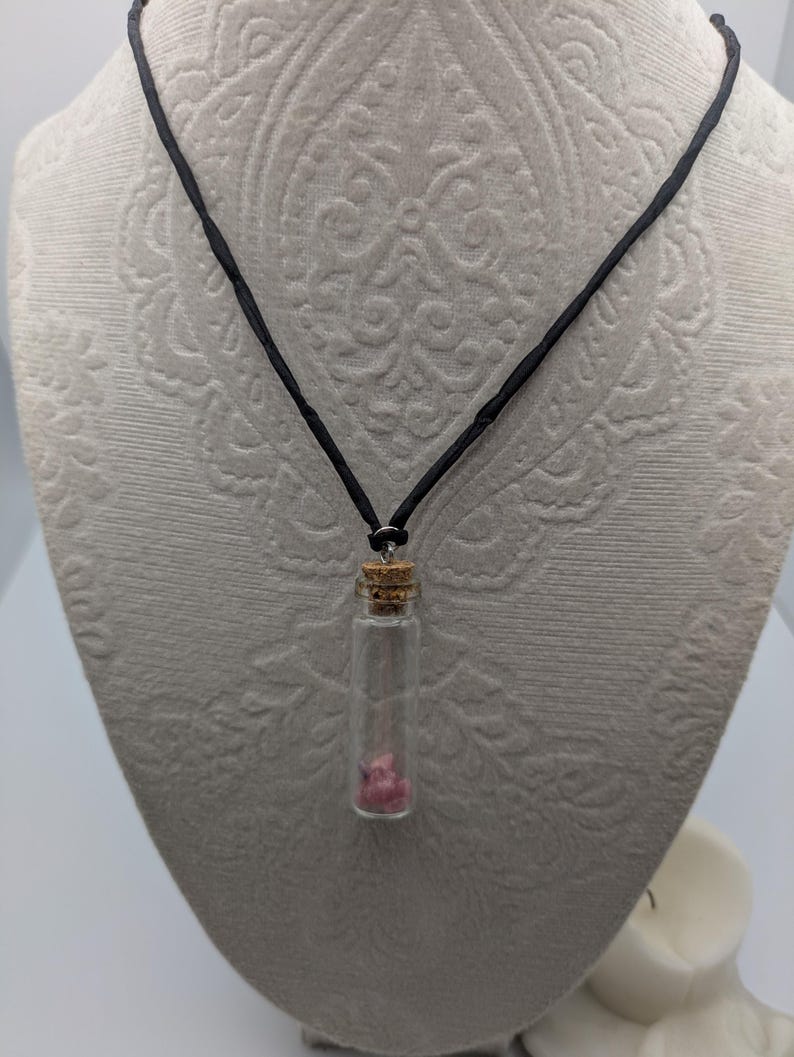 Puede incluir: Un collar de cord&oacute;n negro con un peque&ntilde;o colgante de frasco de vidrio que contiene cristales rosas. El frasco tiene un tap&oacute;n de corcho.