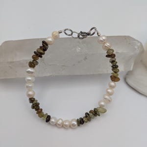 K&ouml;nnte beinhalten: Ein Armband aus wei&szlig;en Perlen und gr&uuml;nen und braunen Edelsteinperlen. Das Armband hat einen silbernen Verschluss.